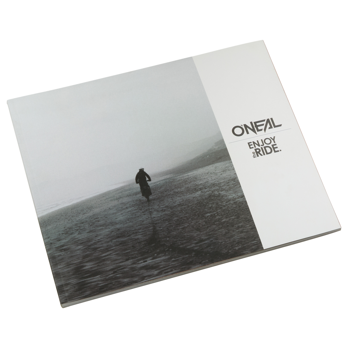 Il libro chiuso O'NEAL ENJOY THE RIDE mostra una figura solitaria su una spiaggia nebbiosa che evoca emozioni. Il logo O'NEAL e le parole "ENJOY THE RIDE." in nero su bianco sono visibili sul lato destro. Marchio: O'NEAL. Prodotto: LIBRO ENJOY THE RIDE di O'NEAL (BAMBINO IN OMAGGIO).