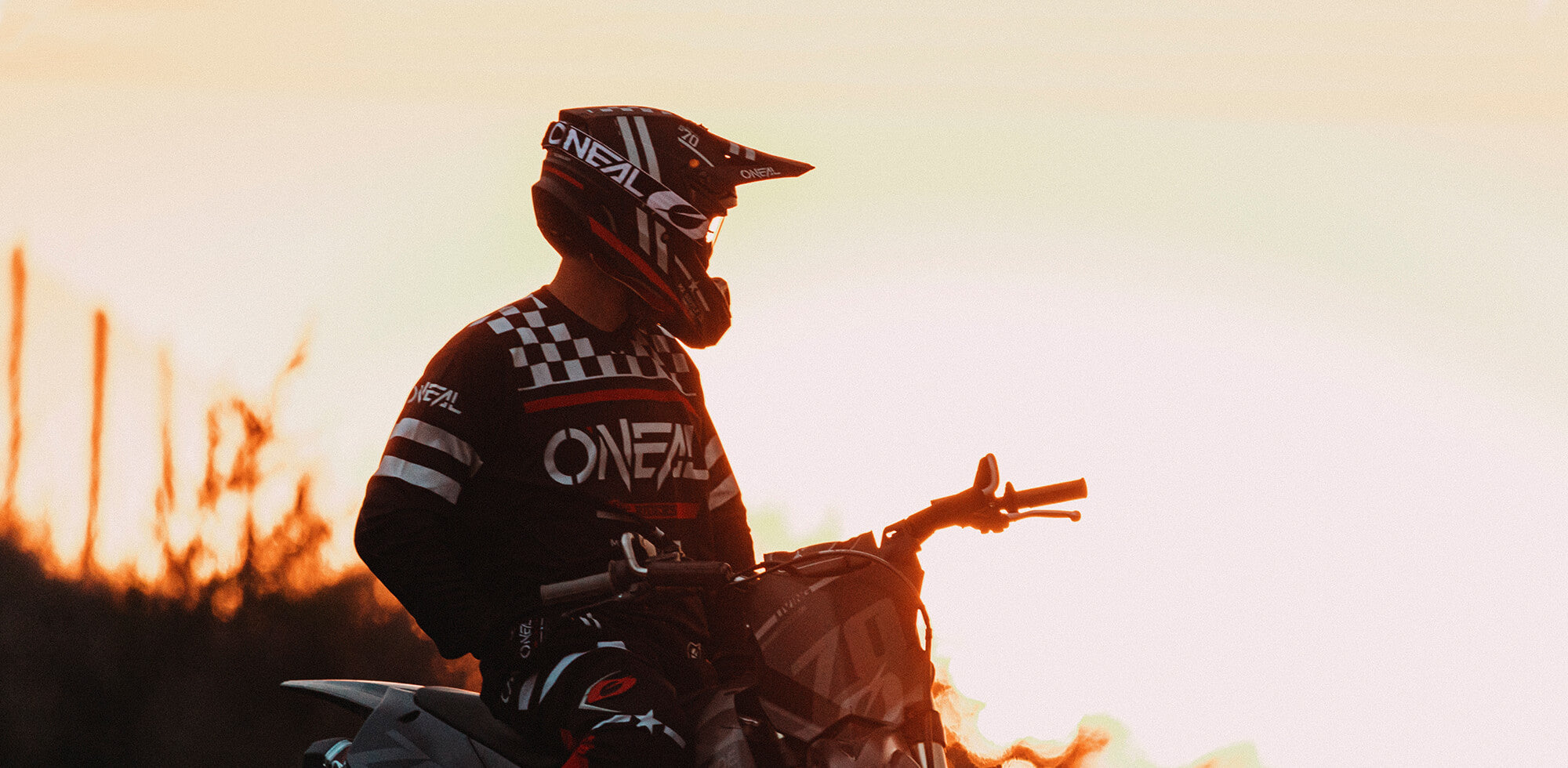 Ein Motocrossfahrer mit Helm und voller Montur sitzt auf einem Dirtbike vor dem Hintergrund eines leuchtenden Sonnenuntergangs. Der Fahrer blickt zur Seite und der Himmel zeigt Schattierungen von Orange und Gelb. Die Szene weckt ein Gefühl von Abenteuer und Freiheit.