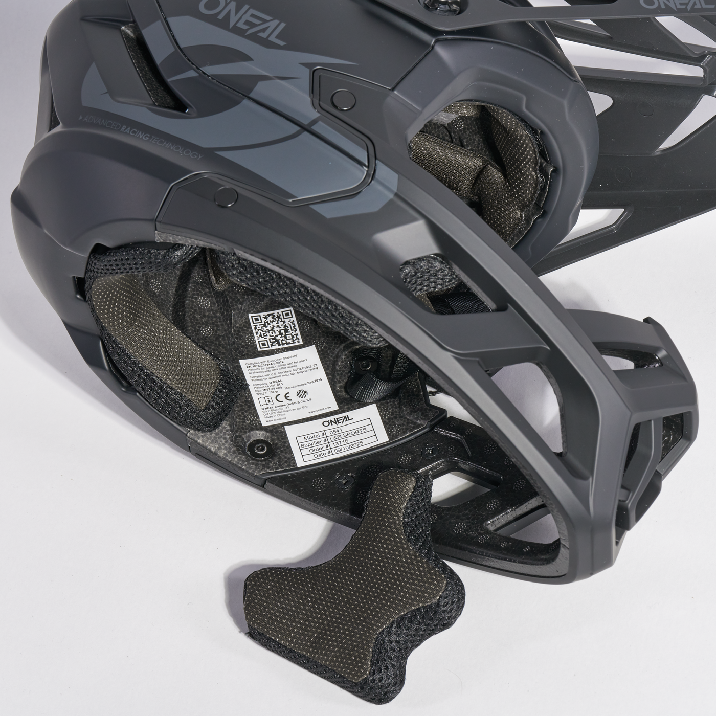 Nahaufnahme eines schwarz-grauen O'Neal-Fahrradhelms, die die Innenpolsterung, den QR-Code-Aufkleber und die strukturellen Details des Kinnschutzes zeigt. Der Helm ruht auf einer weißen Fläche.