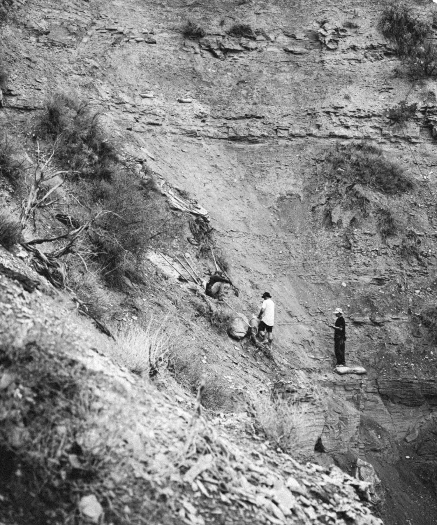 Drei Personen stehen an einem felsigen, abschüssigen Hang und untersuchen den Boden und die Felsen. Auf dem unebenen Gelände wächst eine spärliche Vegetation, und der Hintergrund wird von geschichteten Felsformationen gebildet.