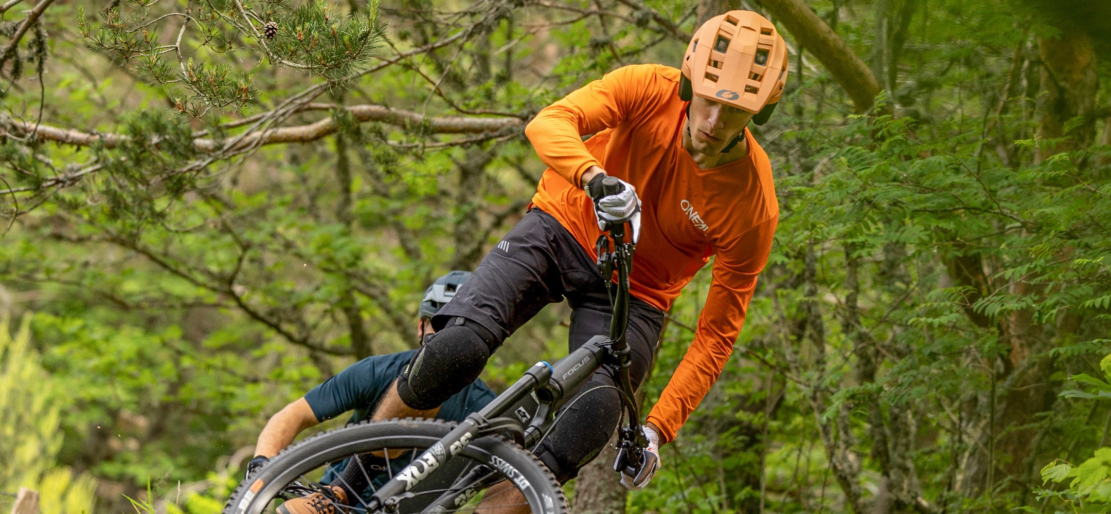 Eine Person mit orangefarbenem Helm und T-Shirt fährt mit einem Mountainbike durch einen üppigen, grünen Wald und legt sich konzentriert und entschlossen in eine Kurve. Hinter ihr ist ein weiterer Radfahrer teilweise zu sehen.