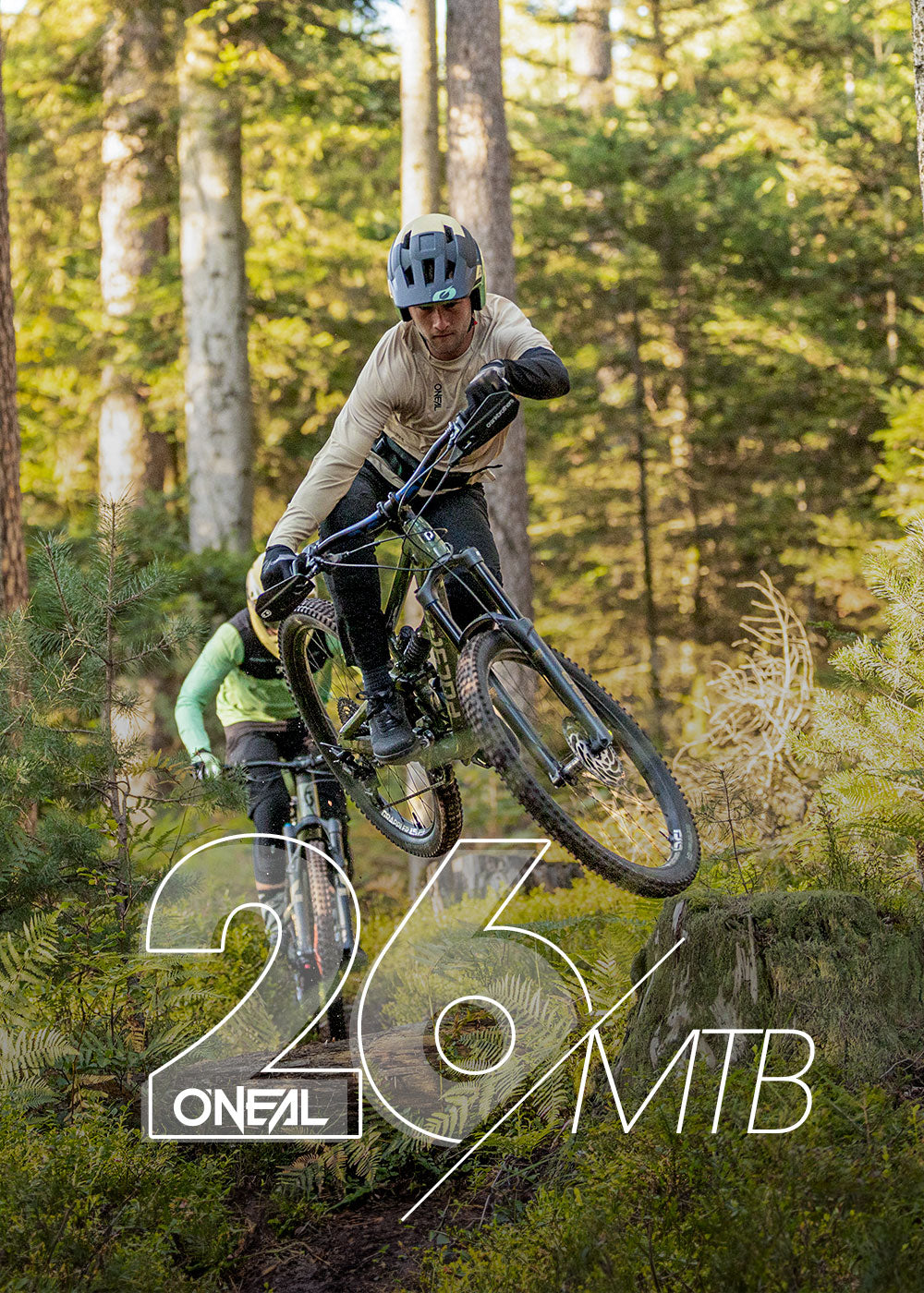 Ein Mountainbiker macht einen Sprung auf einem Waldweg, ein anderer Radfahrer folgt ihm. Das Sonnenlicht fällt durch die hohen Bäume. Der überlagerte Text lautet "26 O'NEAL MTB.