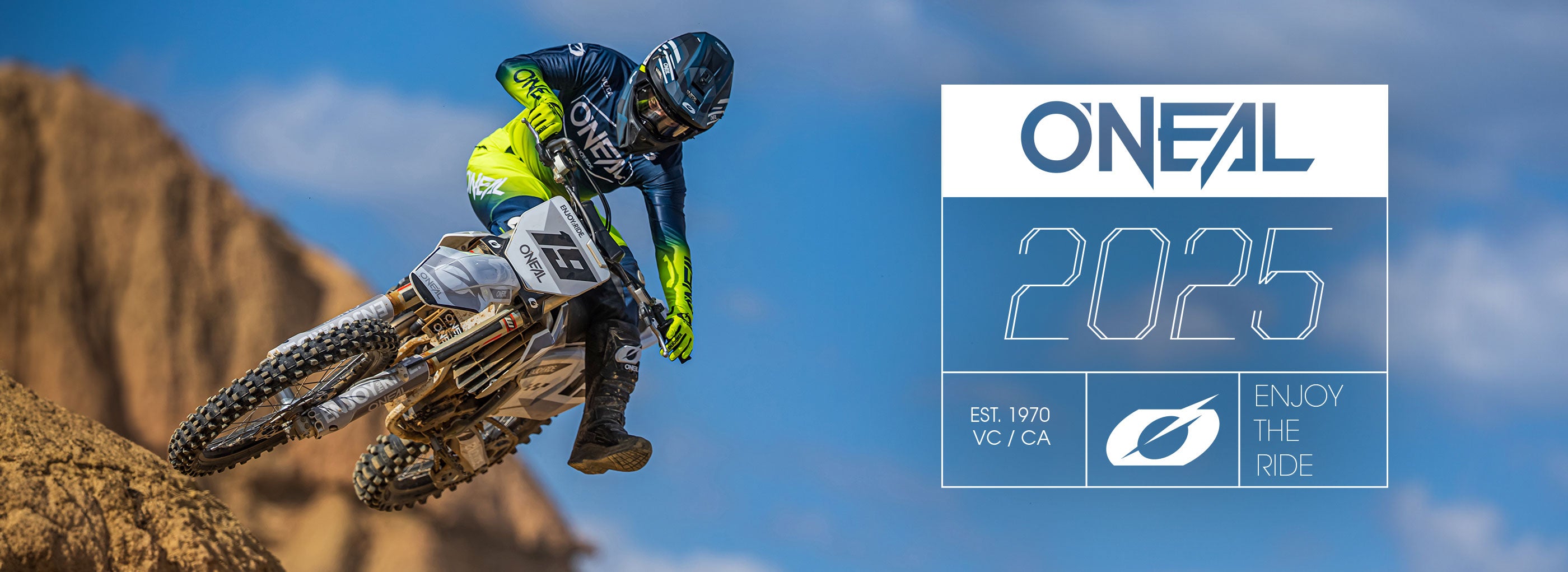 Ein Motocross-Fahrer in blauer und neongelber Ausrüstung springt unter blauem Himmel mit seinem Dirtbike von einem felsigen Hügel. Daneben steht der Text: „O'Neal 2025. Gegründet 1970 VC/CA. Viel Spaß beim Fahren.“.