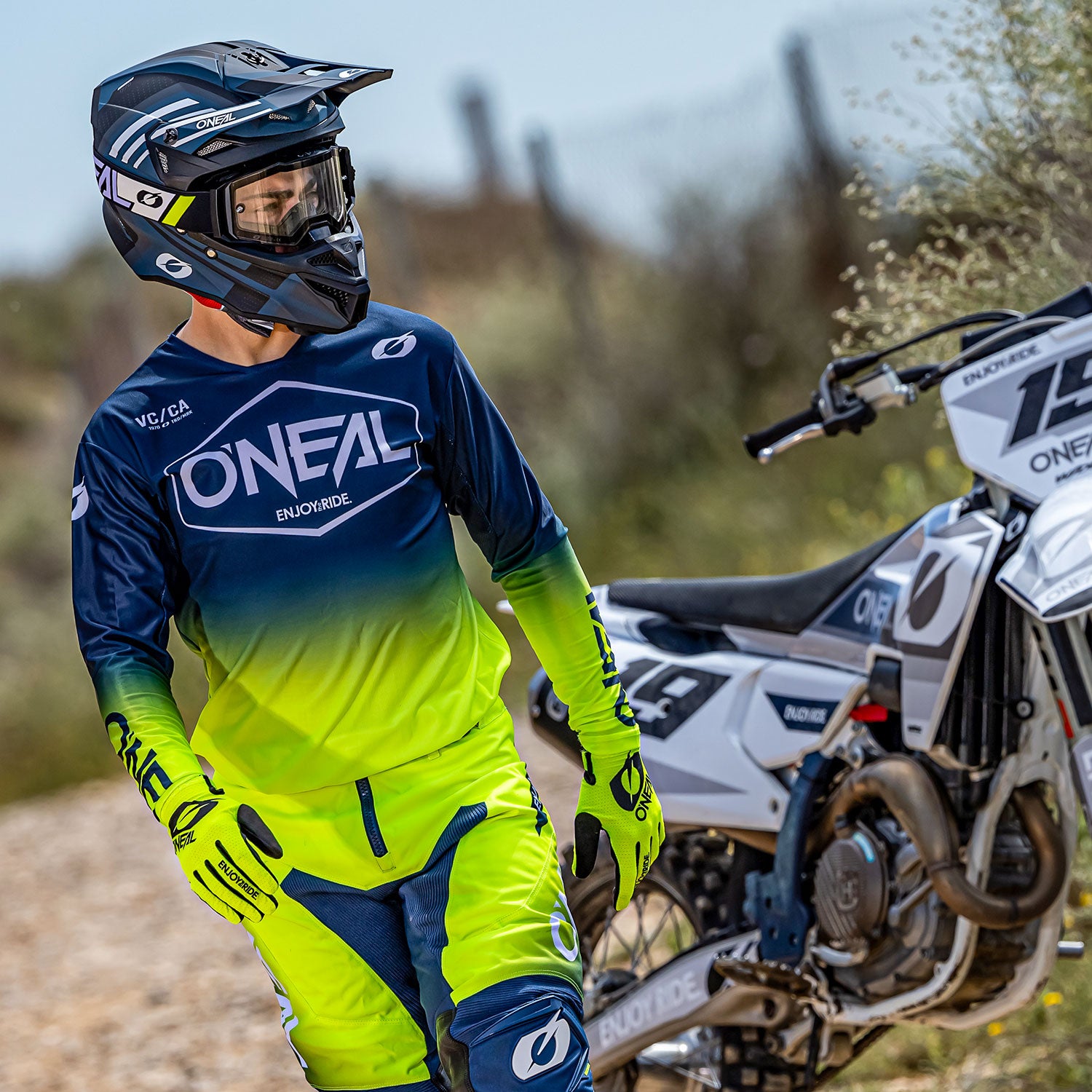 Ein Dirtbike-Fahrer steht neben einem Motorrad. Er trägt ein blau-gelbes O'Neal Enduro-Trikot und passende Hosen sowie einen schwarzen Helm. Im Hintergrund sind ein Feldweg und ein Zaun zu sehen. Der Fahrer blickt zur Seite.
