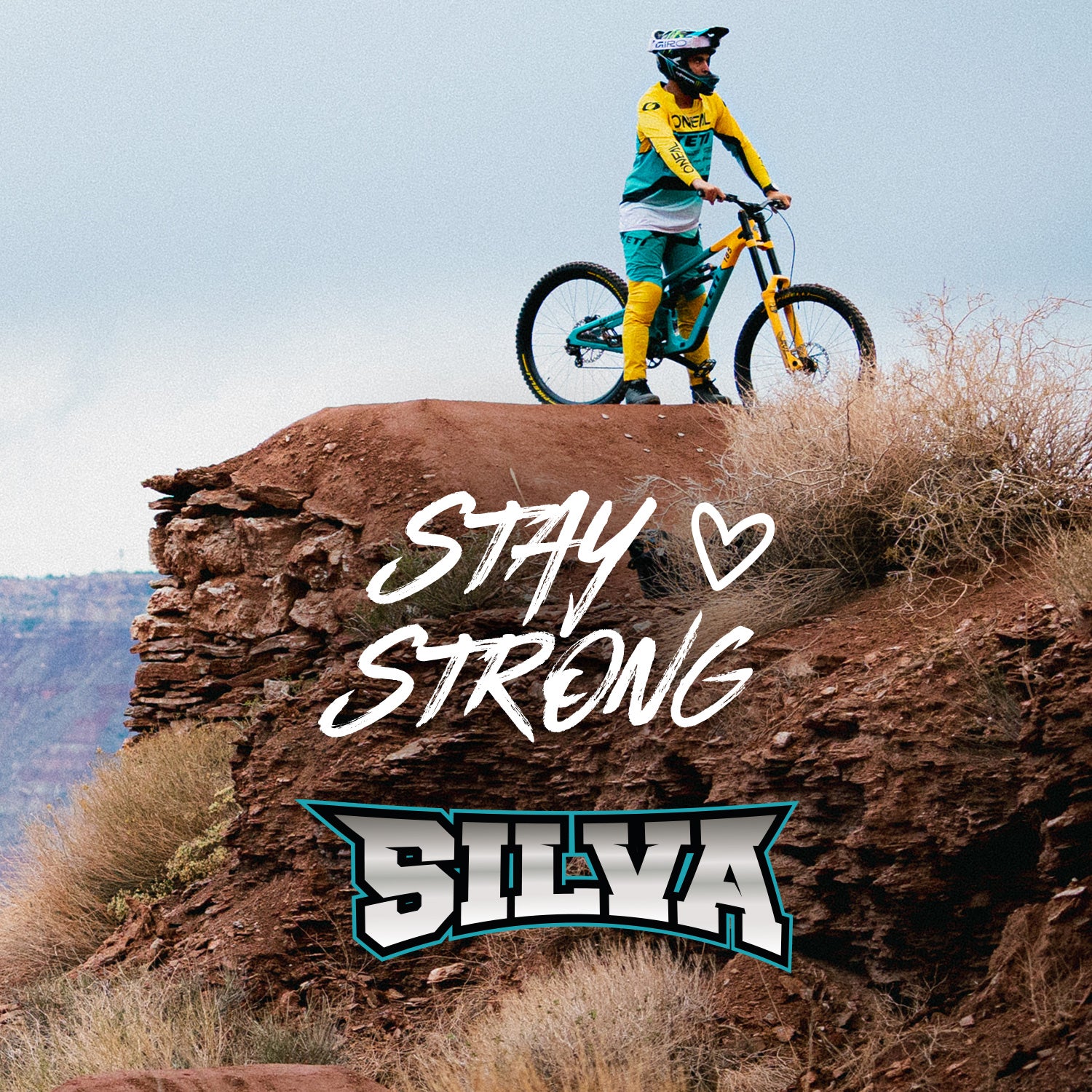 Ein Mountainbiker in gelber und blaugrüner Kleidung steht mit seinem Fahrrad an einem felsigen Klippenrand. Die Worte "Stay Strong" und "Silva" erscheinen über dem Bild, mit einem Herzsymbol zwischen den Worten.
