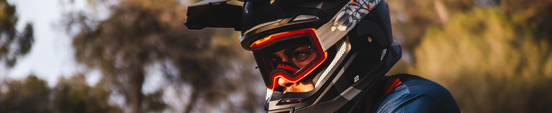 Eine Person, die einen schwarzen Helm mit Visier und eine rote Schutzbrille trägt, befindet sich im Freien, möglicherweise auf einem Mountainbike oder Motorrad, wobei die Bäume im Hintergrund verschwimmen.