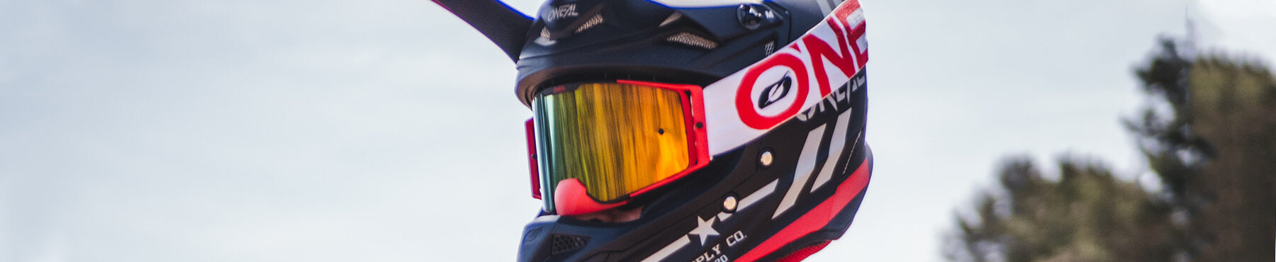 Person, die einen schwarz-roten Motocross-Helm mit reflektierendem Visier trägt, im Freien vor einem hellen Himmel und verschwommenen Bäumen im Hintergrund.