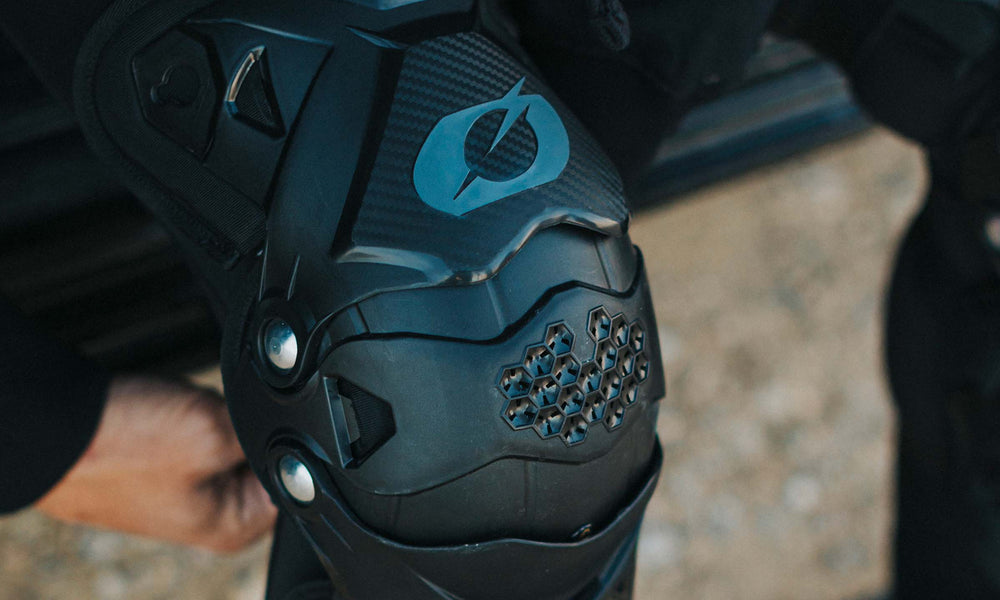 Nahaufnahme einer Person, die einen schwarzen gepanzerten Knieschoner mit blauem Logo trägt. Der Knieschoner hat auf der Vorderseite ein strukturiertes, sechseckiges Design und Metallnieten, was an Schutzausrüstung für Sport oder Motorradfahren erinnert.