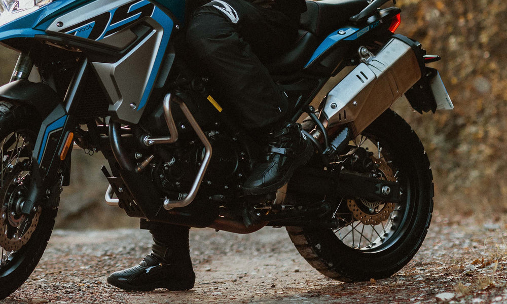 Person auf einem blauen Motorrad mit schwarzer Ausrüstung, geparkt auf einem Schotterweg, umgeben von trockenem Laub.