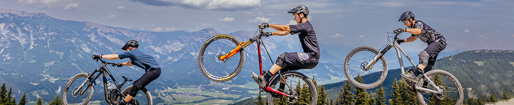 Drei Mountainbiker fahren einen Bergrücken entlang, mit malerischer Aussicht auf Berge und Täler im Hintergrund. Sie fahren in einer Reihe, wobei der mittlere Biker einen Wheelie macht. Der Himmel ist teilweise bewölkt.