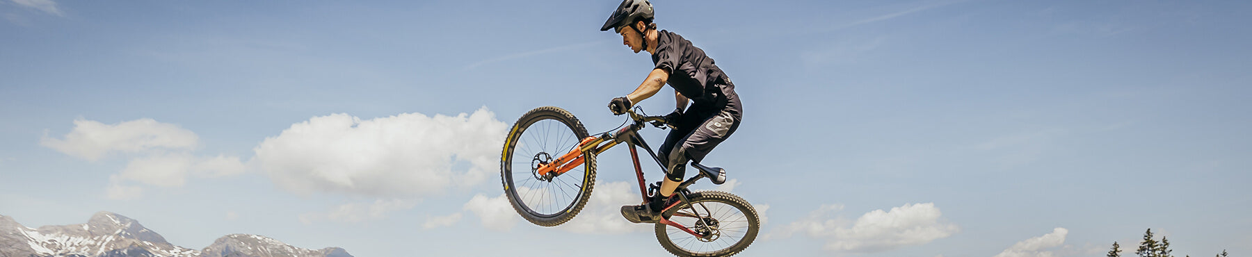 Person, die einen Stunt in der Luft auf einem Mountainbike ausführt und dabei einen Helm und Schutzausrüstung trägt. Im Hintergrund ist ein klarer blauer Himmel mit ein paar Wolken und in der Ferne sind Berge zu sehen.