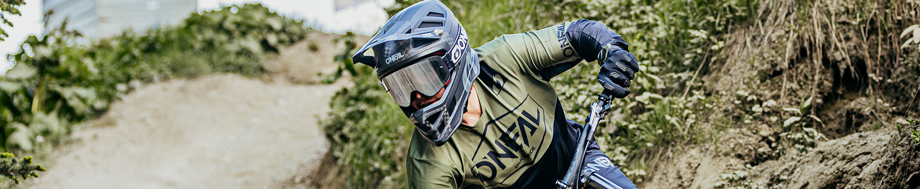 Ein Mountainbiker mit Helm und Schutzbrille fährt einen Feldweg hinunter. Der Weg ist von Grünflächen und Hängen umgeben.