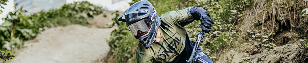 Ein Mountainbiker mit Helm und grünem Trikot befährt einen von Vegetation umgebenen Feldweg und konzentriert sich auf den vor ihm liegenden Weg.