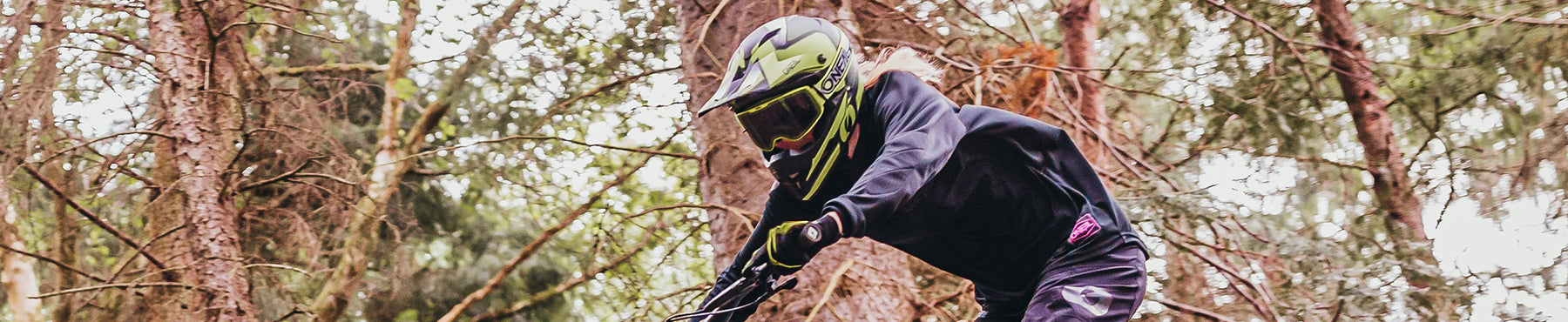Ein Mountainbiker mit grünem Helm fährt durch einen Waldweg. Die Person ist konzentriert und nach vorne gebeugt und navigiert zwischen hohen Bäumen hindurch. Die Szene ist dynamisch und actionreich.