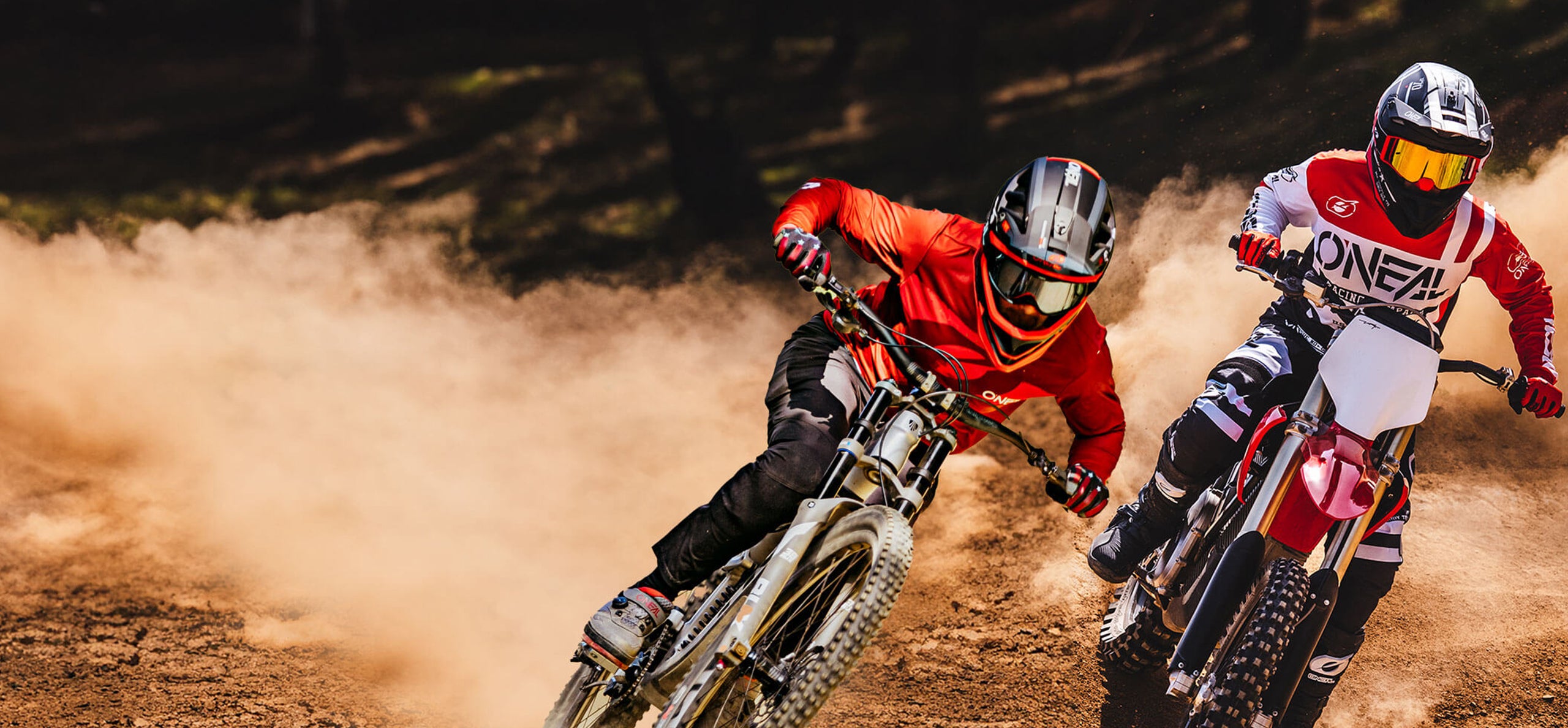 Zwei Motocross-Fahrer in roter Montur rasen auf einer unbefestigten Strecke und wirbeln Staub auf, während sie im hellen Sonnenlicht um eine Kurve rasen.