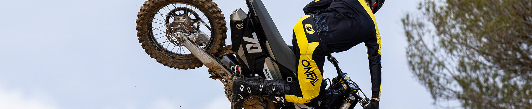 Ein Motocross-Fahrer in gelb-schwarzer Ausrüstung führt vor dem Hintergrund eines bewölkten Himmels einen Stunt in der Luft auf einem Dirtbike aus.