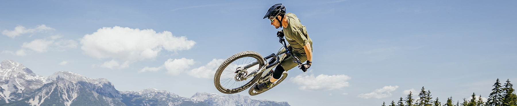 Eine Person mit Helm führt auf einem Mountainbike vor dem Hintergrund eines klaren blauen Himmels und schneebedeckter Berggipfel einen Hochsprung in der Luft aus.
