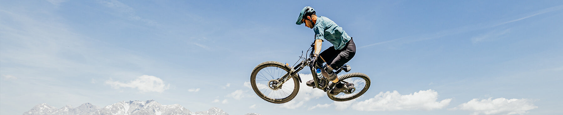 Eine Person in einem blauen Hemd und Helm fährt mit einem Mountainbike durch die Luft vor einem klaren blauen Himmel. In der Ferne sind darunter Berge zu sehen.
