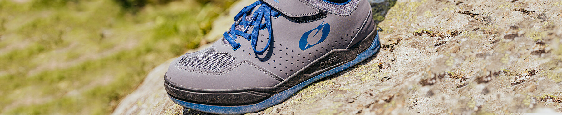 Ein grau-blauer Schuh mit Schnürsenkeln liegt draußen auf einer strukturierten Felsoberfläche. Der Schuh verfügt über Belüftungslöcher und ein stilisiertes Logo an der Seite. Sonnenlicht hebt seine Details und die natürliche Umgebung hervor.
