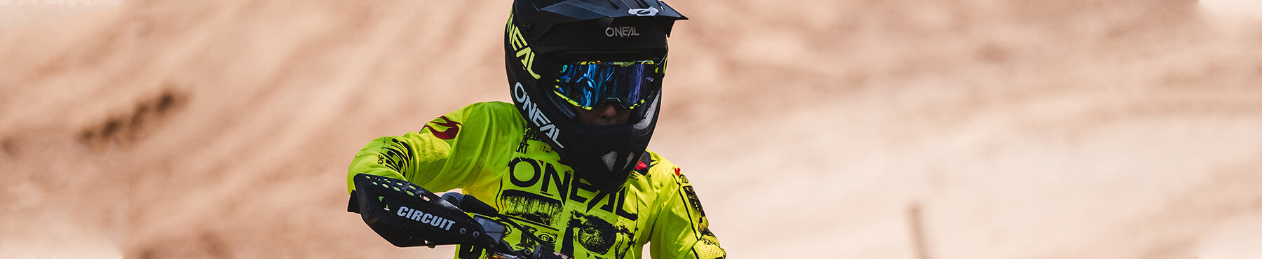 Ein Motocrossfahrer in einem leuchtend gelben Trikot und einem schwarzen Helm mit getönter Schutzbrille fährt mit einem Dirtbike auf einer Sandpiste. Der Hintergrund ist unscharf, um die Bewegung hervorzuheben.