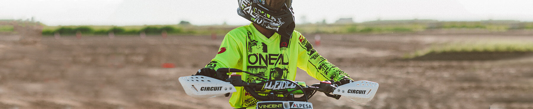 Ein Motocross-Fahrer in neongrüner Ausrüstung und Helm steht auf einem Dirtbike in einem sandigen Gelände, mit verschwommener Naturlandschaft im Hintergrund.