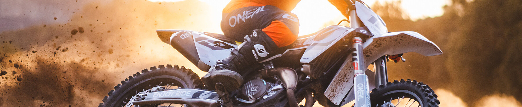 Ein Motocrossfahrer in orange-schwarzer Ausrüstung fährt eine scharfe Kurve auf einer unbefestigten Piste und hinterlässt dabei eine Staubwolke. Im Hintergrund geht die Sonne unter und taucht die Szene in einen warmen Schein.