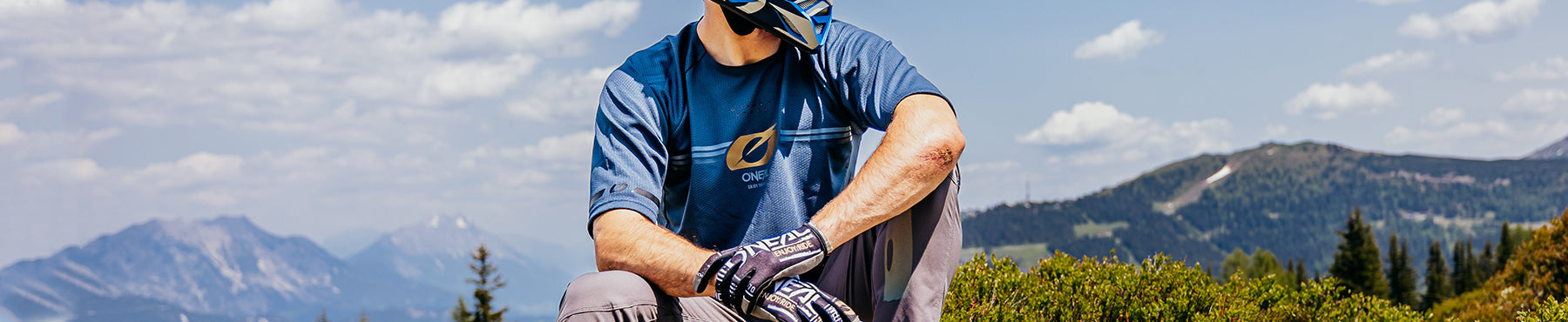 Eine Person mit blauem Helm, Schutzbrille, blauem Hemd und gemusterten Handschuhen sitzt im Freien. Sie ist von bergigem Gelände unter einem teilweise bewölkten Himmel umgeben.