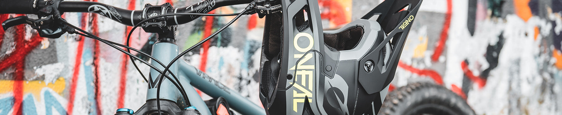 Nahaufnahme eines Mountainbikes, das vor einer mit Graffiti bedeckten Wand abgestellt ist. Der Fokus liegt auf dem vorderen Lenker und der Federung des Fahrrads, an dessen Lenker ein schwarz-grauer Helm hängt.