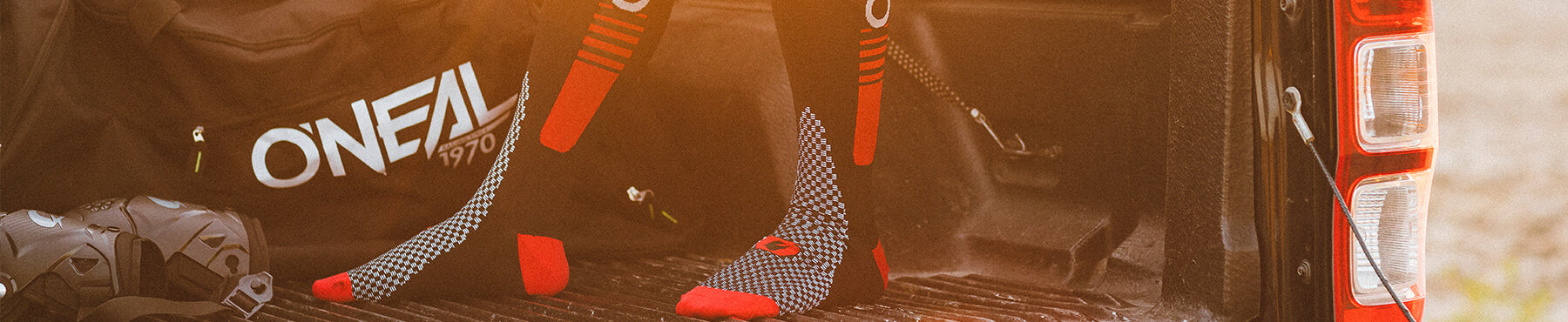 Zwei rot-schwarze Socken liegen auf einer Ladefläche neben einer schwarzen Tasche mit der Aufschrift „ONEAL“ und einem Helm. Der Hintergrund ist sanft verschwommen, in warmes Licht getaucht.
