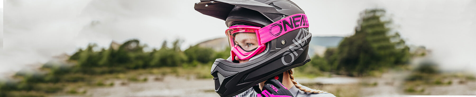 Person, die einen schwarz-rosa Motocross-Helm mit Visier und Schutzbrille trägt, steht im Freien mit verschwommenem Grün im Hintergrund.