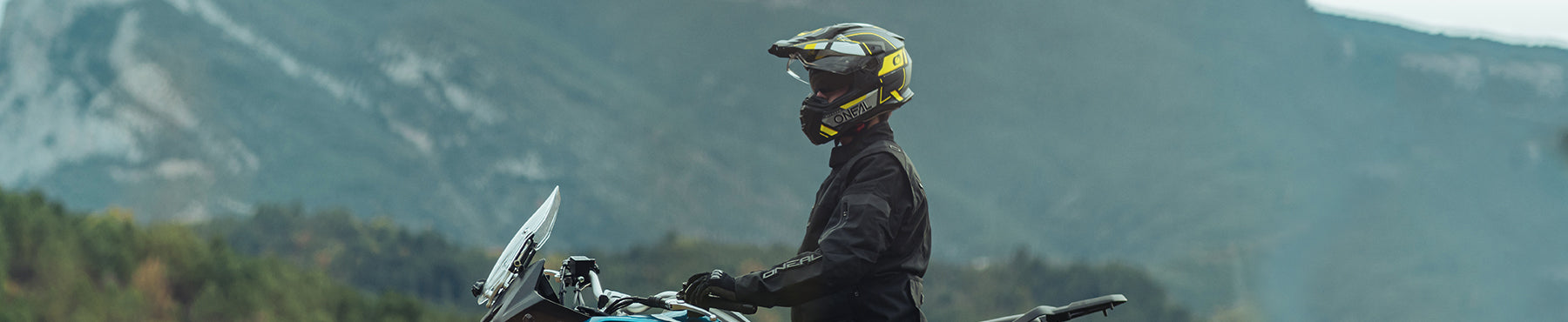 Motorradfahrer in schwarzer Montur und Helm sitzt auf einem Motorrad mit Bergen im Hintergrund. Der Fahrer blickt nach links und ist bereit, durch die malerische Landschaft zu fahren.