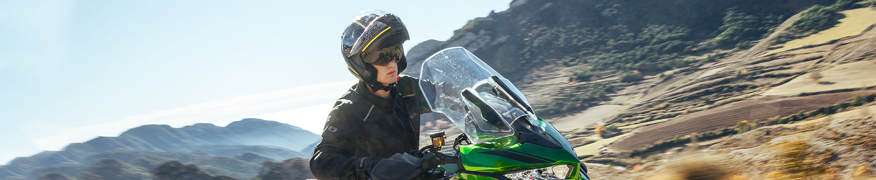 Eine Person mit Helm und schwarzer Jacke fährt auf einem grünen Motorrad durch eine Berglandschaft mit klarem Himmel und zerklüftetem Gelände im Hintergrund.