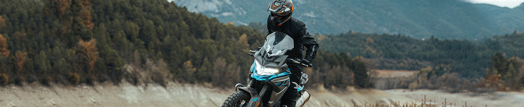 Eine Person in Motorradmontur fährt auf einem Motorrad einen schroffen Pfad entlang, im Hintergrund sind Berge und dichter Wald zu sehen. Der Himmel ist bedeckt, was die abenteuerliche Atmosphäre noch verstärkt.