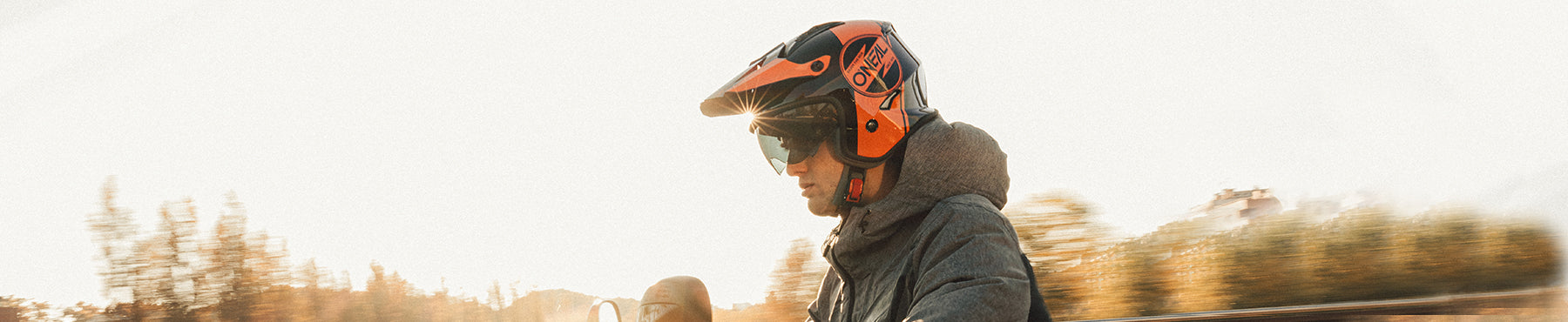 Eine Person mit Helm und Schutzbrille fährt ein Geländefahrzeug vor einem unscharfen Hintergrund aus Bäumen und Himmel, was Bewegung und eine Umgebung im Freien suggeriert.