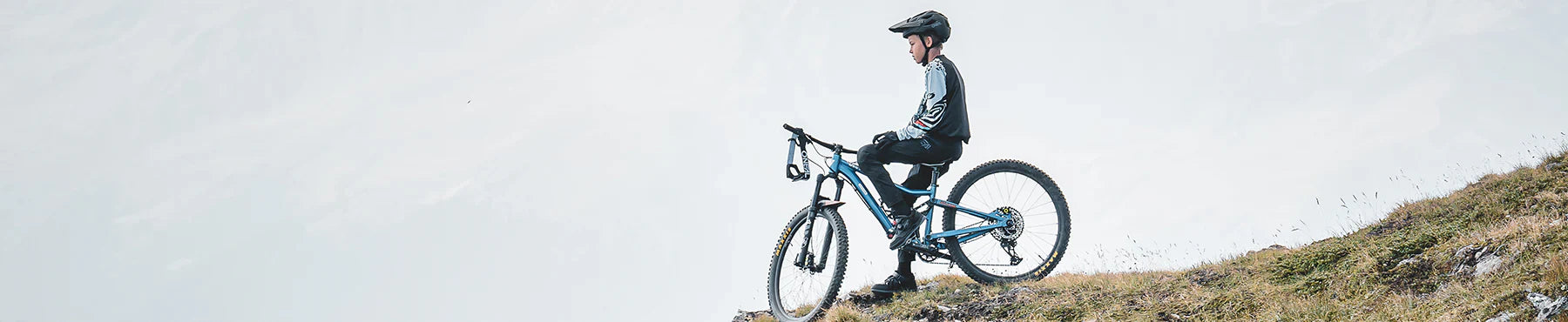 Eine Person in Radfahrerkleidung steht neben einem Mountainbike auf einer grasbewachsenen Hügelkuppe und blickt bei bedecktem Himmel in die dunstige Ferne.
