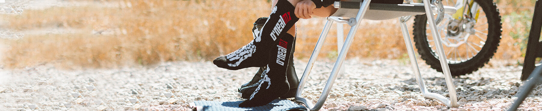 Eine Person in Motocross-Stiefeln sitzt auf einem Klappstuhl neben einem Dirtbike auf einer Schotterfläche. Sie richtet die Stiefel, im Hintergrund ist hohes Gras zu sehen.