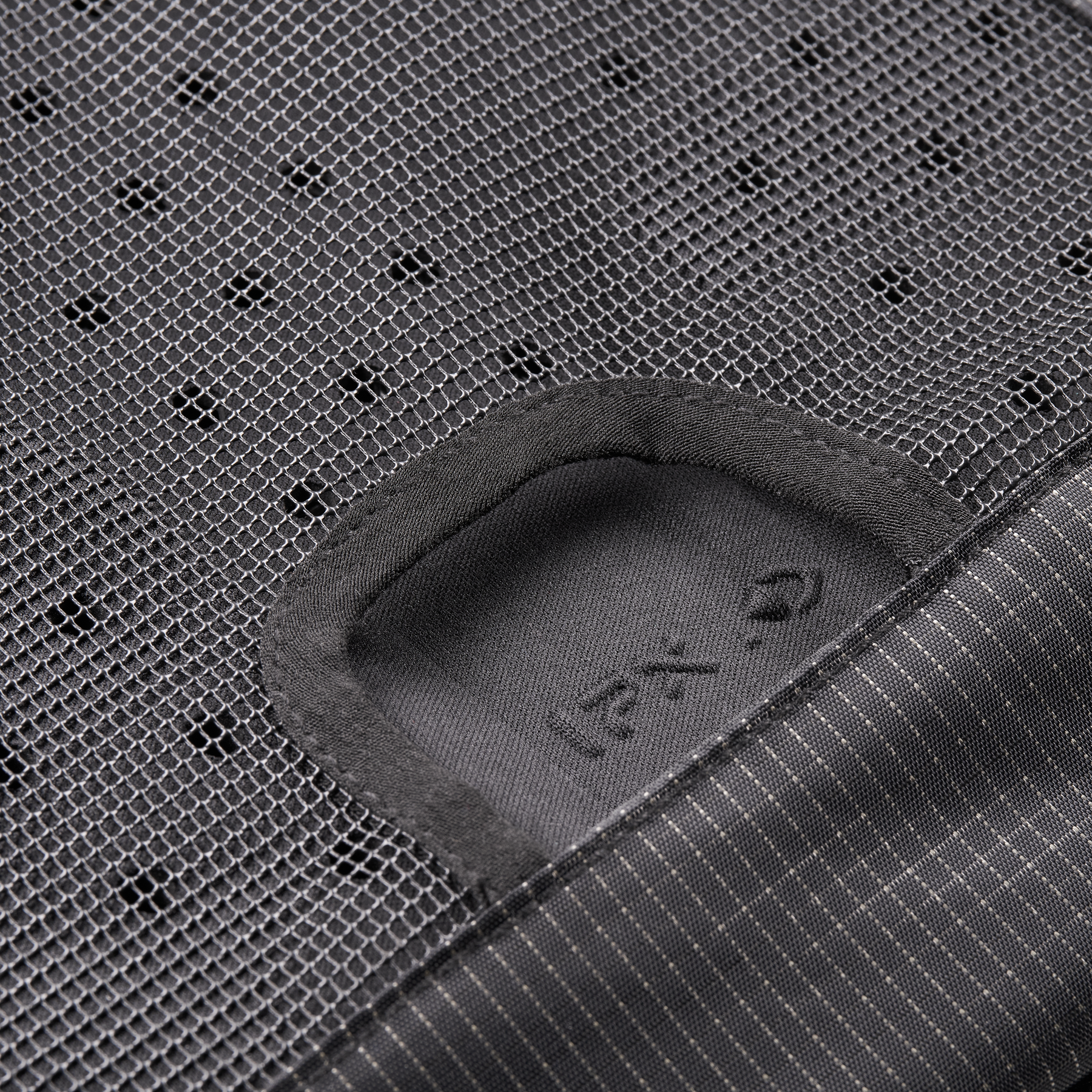 Nahaufnahme des schwarzen Mesh-Gewebes des O'NEAL Europe JUNCTION MTB Protektorrucksacks, die einen rechteckigen Ausschnitt mit einem weichen, dunklen Innenaufnäher mit dezentem Text und Symbolen zeigt; unten rechts erscheint der Nadelstreifenstoff.