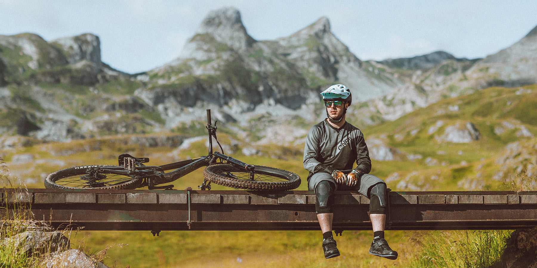 Ein Radfahrer mit Helm und Schutzausrüstung sitzt auf einer Holzbrücke in einer Berglandschaft. Sein Fahrrad liegt seitlich neben ihm auf der Brücke. Die Szene zeigt schroffe, grüne Berge unter einem teilweise bewölkten Himmel.