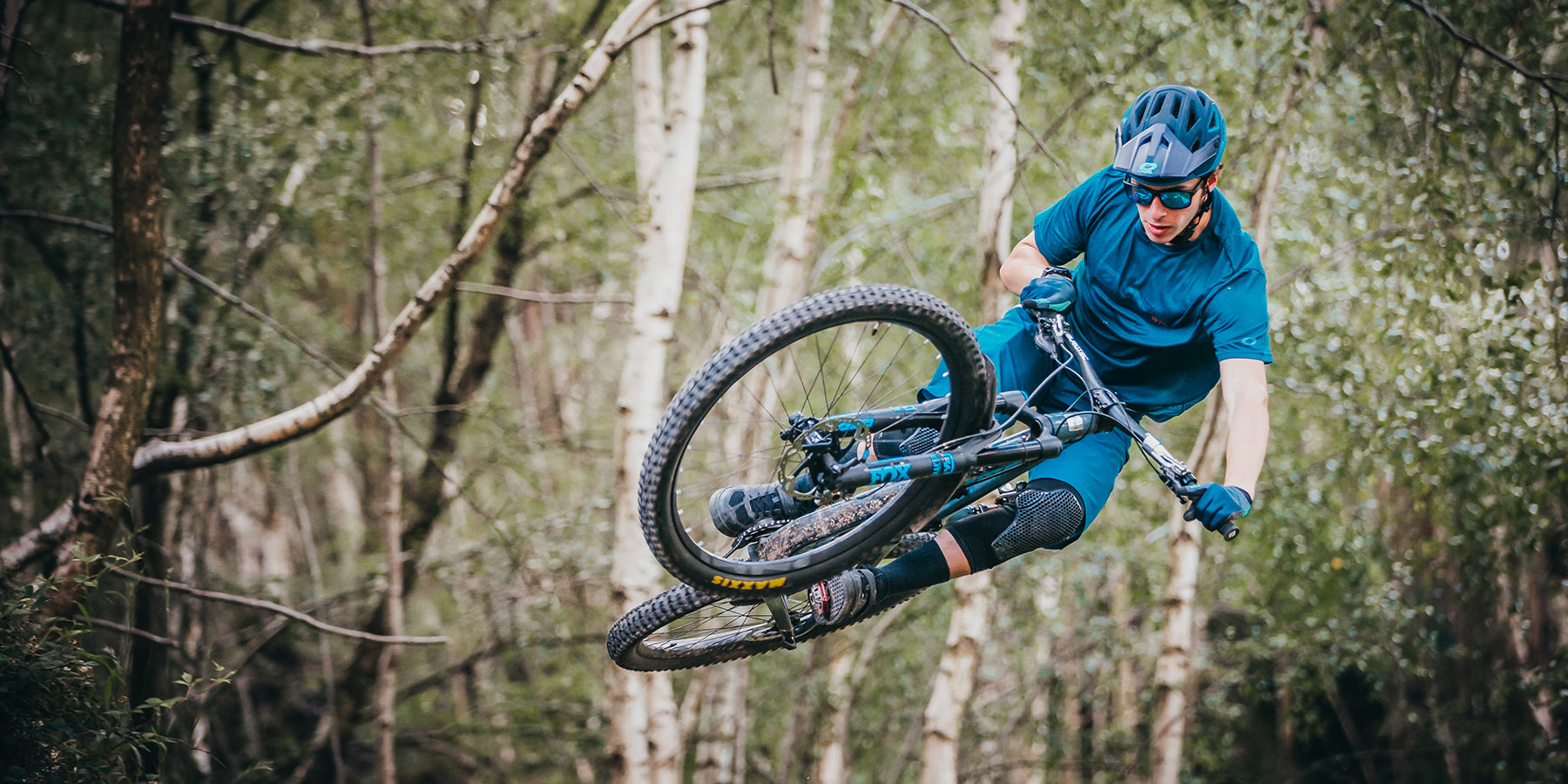 Ein Radfahrer in blauer Ausrüstung führt in einem Waldgebiet mit Birken einen Lufttrick auf einem Mountainbike vor. Der Radfahrer ist seitlich geneigt und zeigt geschickte Kontrolle und Balance. Der Hintergrund ist voller Grün, was die abenteuerliche Szene noch verstärkt.
