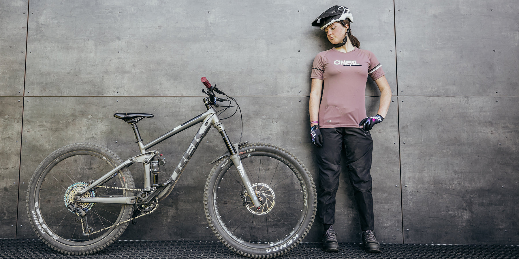 Eine Person in einem rosa O'Neal-Trikot, einer schwarzen Hose, einem Helm und Handschuhen steht an einer Betonwand und blickt auf ein Mountainbike neben ihr. Das Fahrrad mit dicken Reifen und einem stabilen Rahmen steht auf einem Boden aus Metallgitter.
