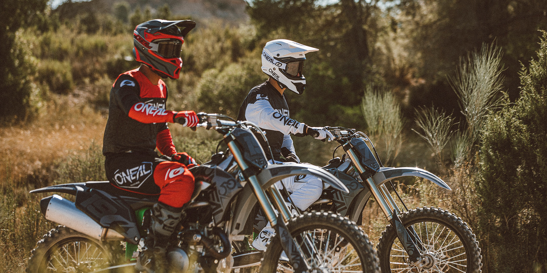 Zwei Motocrossfahrer sind auf ihren Dirtbikes im Freien unterwegs. Sie tragen die volle Montur, einschließlich Helme, Handschuhe und Motorradanzüge. Ein Fahrer trägt rot-schwarze Montur, der andere weiß-schwarze. Sie scheinen innezuhalten und auf die Strecke zu blicken.