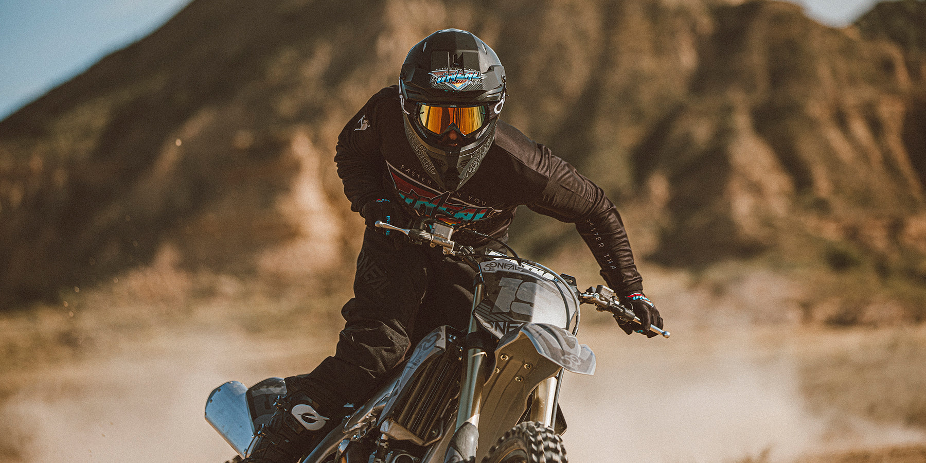 Ein Motocrossfahrer mit schwarzem Helm und Schutzbrille sowie schwarzer Ausrüstung lehnt sich mit seinem Dirtbike in eine Kurve. Der Hinterreifen ist von Staub umgeben, als er von der Strecke abhebt. Felsige Hügel und ein blauer Himmel bilden den Hintergrund.