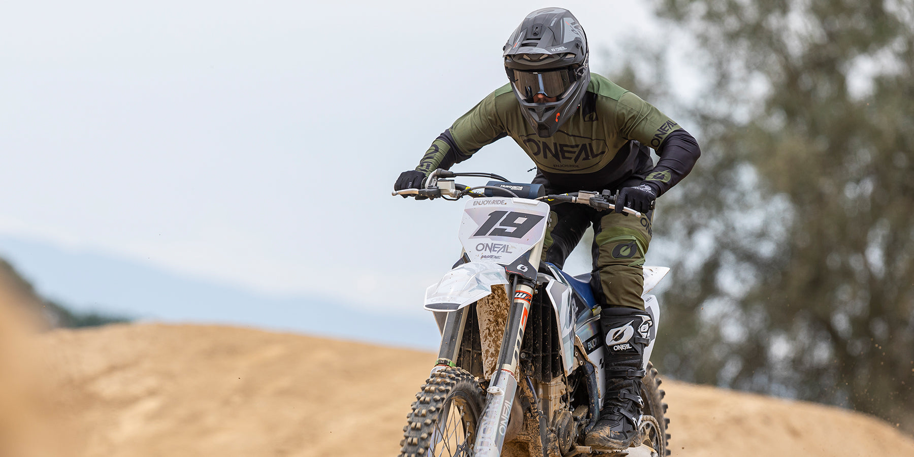 Eine Person mit Helm und Schutzausrüstung fährt mit einem Dirtbike mit der Nummer 19 auf einer Sandpiste. Der Fahrer wirkt konzentriert und hält den Lenker fest umklammert, während die Reifen des Bikes den Staub vom Boden aufwirbeln. Im Hintergrund sind Bäume und ein dunstiger Himmel zu sehen.