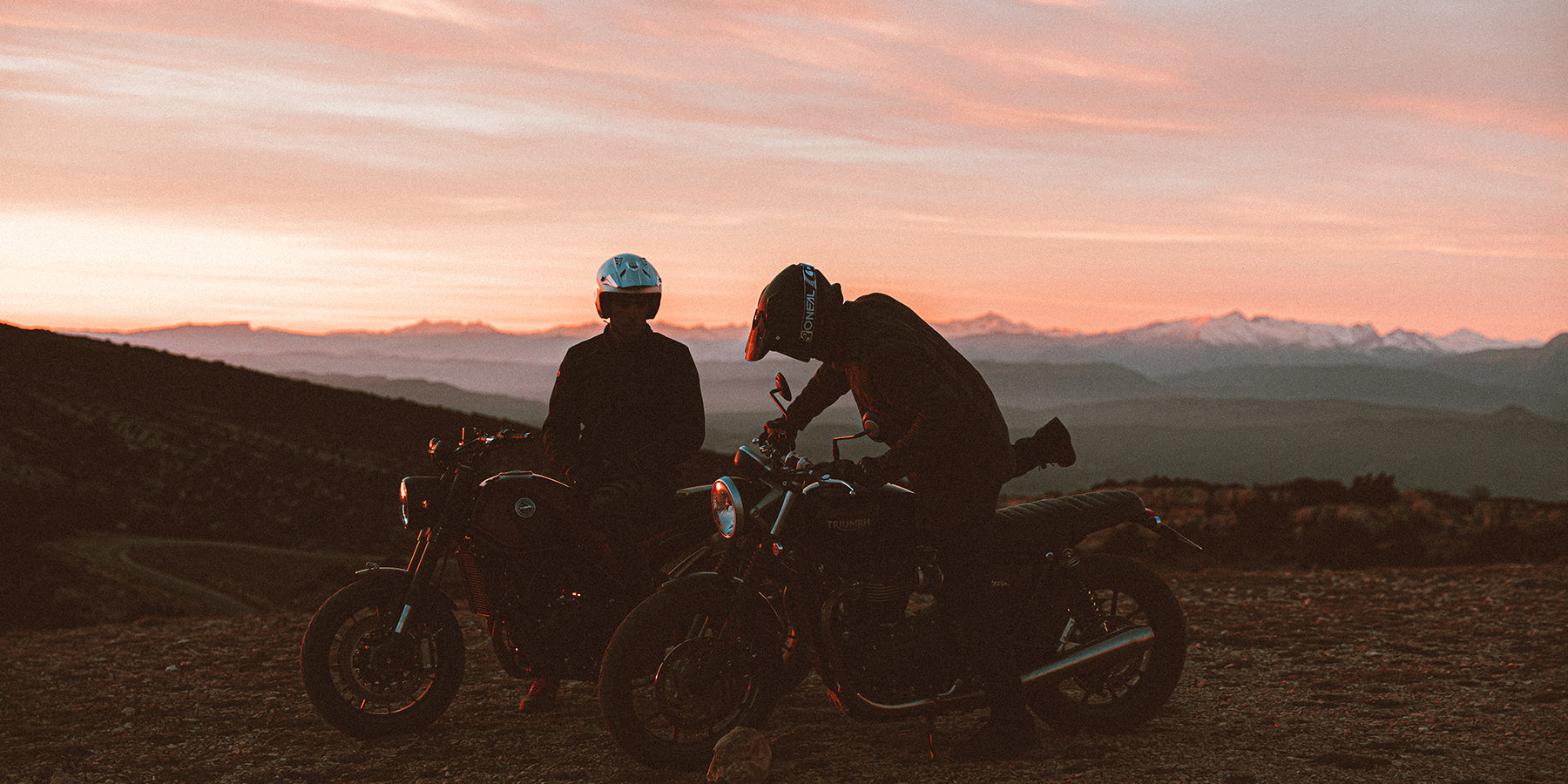 Zwei Motorradfahrer mit Helmen sitzen nebeneinander auf ihren Motorrädern und genießen eine malerische Aussicht bei Sonnenuntergang. Ein Motorradfahrer sitzt aufrecht, während der andere nach vorne gebeugt ist. Schneebedeckte Berge und ein farbenfroher Himmel bilden eine atemberaubende Kulisse.