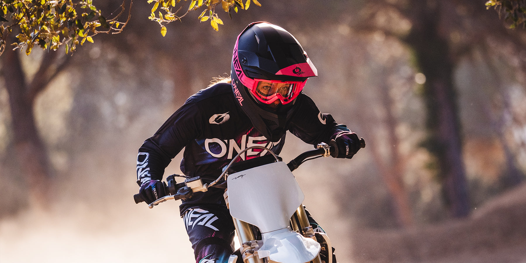 Eine Person mit einem schwarz-rosa Helm, einer Schutzbrille und einem schwarzen Outfit mit dem Wort „ONEAL“ fährt mit einem Dirtbike auf einem staubigen, von Bäumen umgebenen Pfad. Der Fahrer ist konzentriert und hält den Lenker fest umklammert.