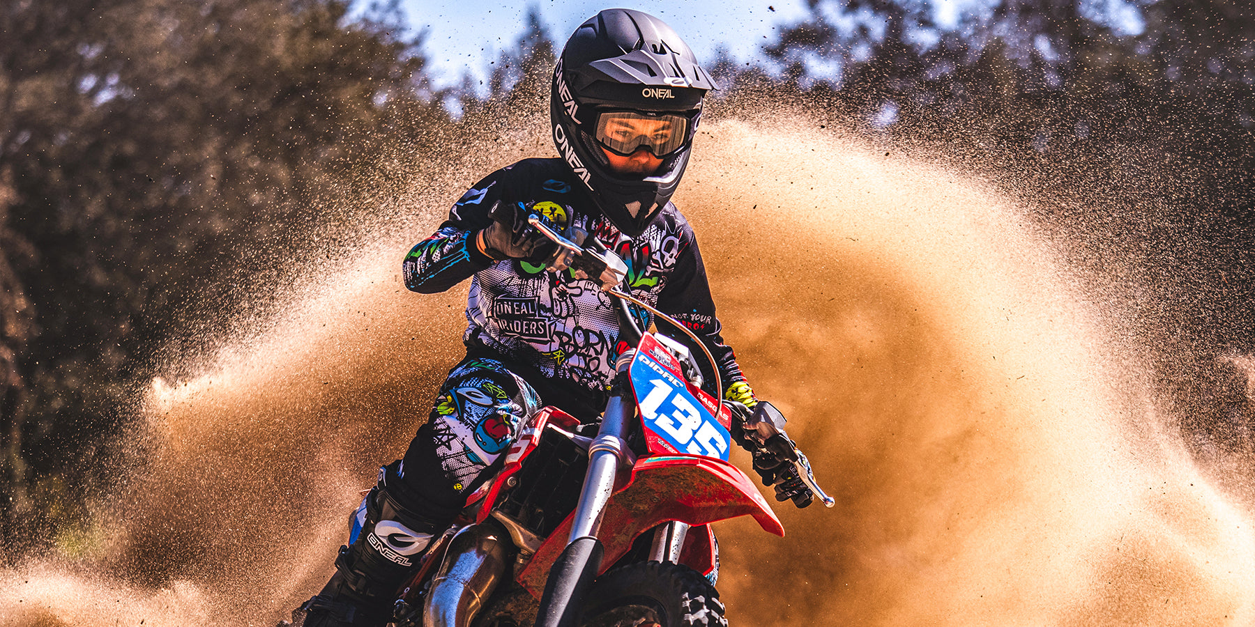 Ein Motocrossfahrer mit Helm und Rennanzug fährt ein rotes Dirtbike und wirbelt dabei eine große Staubwolke hinter sich her. Das Visier des Fahrers reflektiert die Umgebung und die lebhaften Grafiken auf Anzug und Motorrad heben sich vom erdigen Hintergrund ab.