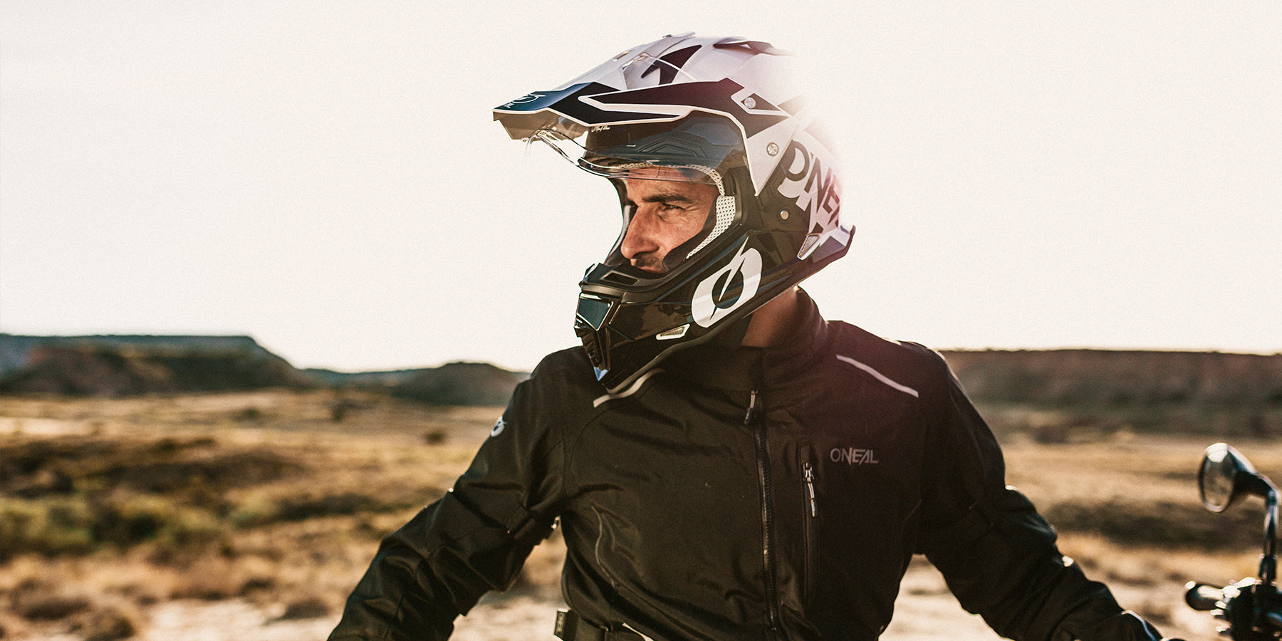 Eine Person, die eine schwarze O'Neal-Motorradjacke und einen weiß-schwarzen Helm trägt, blickt in einer Wüstenlandschaft zur Seite. Die Sonne scheint hell und erhellt die Szene mit einem warmen Schein.