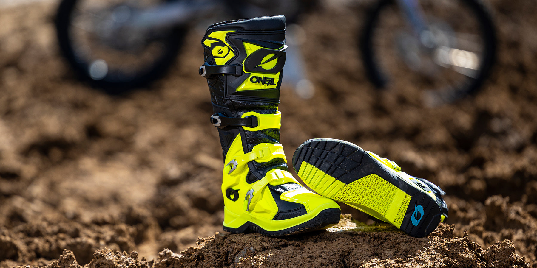 Ein Paar leuchtend gelbe Motocross-Stiefel liegt in einer schlammigen Außenumgebung auf dem Boden. Ein Stiefel steht aufrecht, während der andere auf der Seite liegt und das raue Profil der Sohle zeigt. Im Hintergrund ist ein verschwommenes Motorrad zu sehen.
