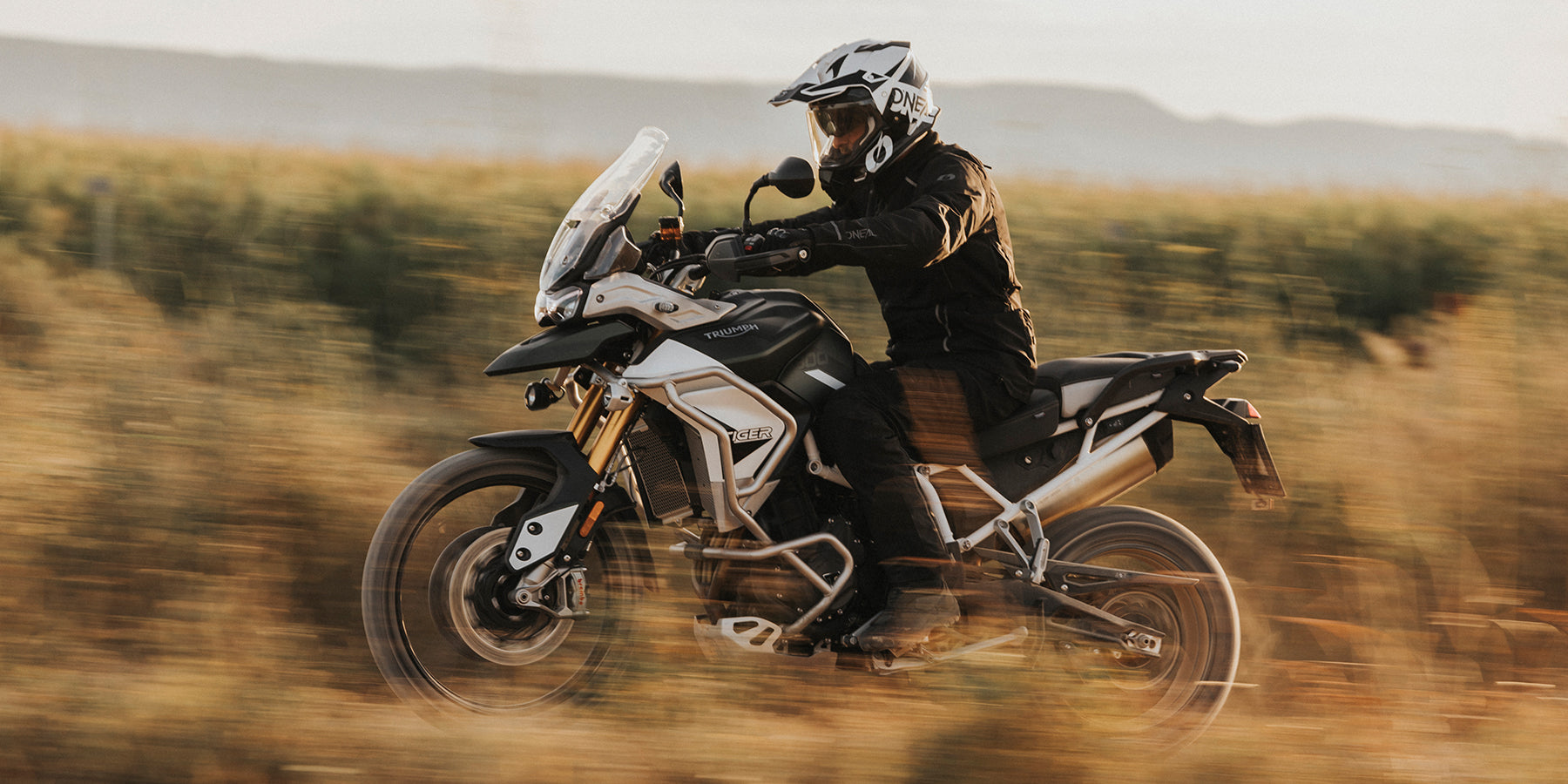 Person, die mit einem weißen und schwarzen Adventure-Motorrad durch eine Wiese fährt und einen weißen Helm und schwarze Schutzkleidung trägt. Der Hintergrund zeigt verschwommenes Grün und einen dunstigen Himmel, was ein Gefühl von Geschwindigkeit und Bewegung vermittelt.