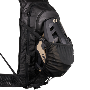 Ein schwarzer O'NEAL JUNCTION MTB Protektorrucksack von O'NEAL Europe mit einer Außentasche aus Mesh, in der ein schwarzer und beiger Fahrradhelm Platz findet, wird vor einem schlichten weißen Hintergrund gezeigt.